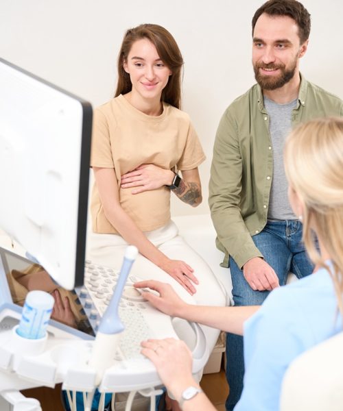 Female caucasian gynecologist looking at young smiling european couple watching result of ultrasound scanning of woman patient pregnant belly on screen in clinic. Concept of pregnancy and paternity